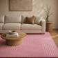 A styled living room scene featuring a large pink area rug with a wavy, curved geometric pattern. The rug is styled in a modern living room setting with a beige fabric sofa, a round wooden coffee table, a side table with a vase of branches, and a neutral-toned wall.