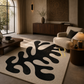 A large modern area rug with a bold black abstract organic pattern on a cream background, styled in a warm, contemporary living room next to a beige armchair and wooden furniture.
