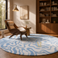 A large round area rug with a wavy abstract pattern in light blue and off-white, styled in a warm, modern living room next to a mid-century modern wooden armchair and a bookshelf.