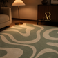 A modern area rug with a wavy, organic abstract pattern in sage green and cream, styled in a warm, contemporary living room setting with a lit floor lamp and a wooden sideboard.