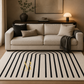 A modern living room scene featuring a large cream-colored area rug with bold black vertical stripes and an arched geometric pattern. The rug is styled in front of a light beige fabric sofa with black and beige throw pillows, a dark wood console table, and a lit table lamp in the background.