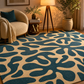A large area rug with an abstract organic pattern in deep teal blue and warm beige, styled in a cozy living room setting with a cream boucle armchair, a floor lamp, and potted plants.
