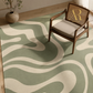 A modern area rug with a wavy abstract pattern in sage green and cream, styled in a room setting with a mid-century modern wooden armchair and a potted plant.
