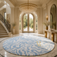 A large round area rug with a light blue and off-white wavy pattern and a gold monogram, displayed in a luxurious, high-ceilinged entryway with marble floors, a crystal chandelier, and a staircase.