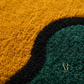Close-up of a textured area rug with a wavy, organic pattern in mustard yellow, black, and deep teal green. A small gold watermark logo for 'AR AROMA RUGS' is visible in the bottom right corner.