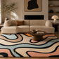 A modern living room scene featuring a large abstract-patterned area rug with flowing wavy lines in beige, black, peach, and light blue. The rug is styled in a warm, contemporary living room with a cream-colored sectional sofa, a round wooden coffee table, and a black fireplace in the background.
