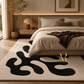 A modern beige area rug with a bold black abstract organic pattern, styled in a warm, contemporary bedroom setting next to a bed with neutral-toned bedding.