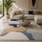 A modern living room scene featuring a large abstract area rug with beige, navy, and slate blue shapes. The rug is styled under a cream boucle sectional sofa, a round coffee table, and a potted palm in a bright, modern interior.