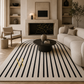 A large area rug with vertical black stripes on a cream background, styled in a modern living room with a beige sofa, black coffee table, and fireplace.