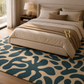 A styled bedroom scene showing a bed with beige bedding and a brown throw blanket, placed in a real interior with a large abstract-pattern area rug in navy and beige, a nightstand, lamp, and window.