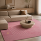 A pink area rug with white wavy line patterns and a gold monogram logo, styled in a modern living room with a beige sectional sofa and a round wooden coffee table.
