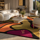 A large area rug with an abstract wavy pattern in orange, yellow, magenta, and black, styled in a modern living room with a cream sofa and marble coffee table.