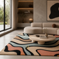 A modern living room scene featuring a large abstract wavy pattern area rug in beige, black, peach, and light blue. The rug is styled in a contemporary living space with a curved beige sofa, a round wooden coffee table, and a concrete wall background.