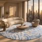 A luxury modern living room featuring a large round area rug with a wavy blue and cream pattern, a curved cream sofa, a matching accent chair, and a round stone coffee table, all set against a backdrop of a city skyline visible through floor-to-ceiling windows at sunset.