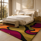 A large area rug with bold abstract wavy patterns in magenta, yellow, orange, olive green, and black, styled in a modern, sunlit bedroom with a cream upholstered bed and matching bench.