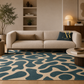 A modern living room scene featuring a beige sofa with a blue and beige abstract patterned area rug underneath. The room has warm neutral walls, a built-in shelf, a lit table lamp, and a potted plant.