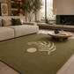 An olive green area rug with a cream palm leaf and sun motif, styled in a modern living room setting with a beige sofa and wooden coffee table.