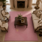 A pink area rug with a modern geometric pattern of concentric arches and fine vertical stripes, styled in a luxurious living room with a fireplace, beige sofas, and a dark wood coffee table.