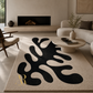 A large rectangular area rug in a modern living room setting. The rug has a warm cream base with a bold, organic black abstract pattern and a small gold monogram logo in the bottom-left corner.