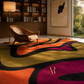 A large area rug with bold abstract wavy patterns in orange, yellow, black, and magenta, styled in a modern living room with a wooden chair, bookshelves, and a window view.
