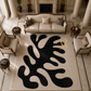 A large modern area rug with an abstract black organic pattern on a cream background, styled in a luxurious, high-ceilinged living room with a sofa, armchairs, and a fireplace.