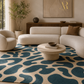 A modern living room scene featuring a large abstract pattern area rug in navy blue and beige, styled with a curved cream sofa, a matching armchair, a low round coffee table, and neutral decor.