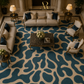 An overhead view of a luxurious living room featuring a large abstract patterned area rug in teal blue and beige, styled with cream sofas, matching armchairs, a dark marble coffee table, and elegant table lamps.