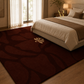 A large rectangular area rug in a warm brown and burgundy abstract pattern with a small beige floral motif, styled in a modern bedroom next to a bed with beige bedding.