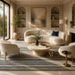 A luxury living room featuring a curved cream bouclé sofa, two matching armchairs, a round stone coffee table, and a striped blue and beige area rug. The room is styled with built-in shelves, a fireplace, and a large abstract painting on the wall.
