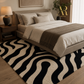 A modern area rug with a bold black and cream wavy zebra-like pattern, styled in a contemporary bedroom setting under a bed with neutral bedding.