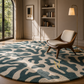 A large round area rug with an abstract organic pattern in teal blue and cream, styled in a modern living room with a mid-century armchair, a floor lamp, and built-in bookshelves.