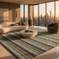 A large area rug with horizontal stripes in muted blue, green, beige, and brown tones, styled in a modern living room with a curved cream sofa, a round stone coffee table, and a dark green armchair, overlooking a city skyline at sunset.