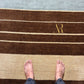 A top-down view of a striped area rug with dark brown, beige, and light blue sections, featuring a gold embroidered monogram 'AR' and bare feet standing on it.