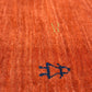 Close-up of a textured orange woven rug with a dark blue embroidered monogram and a faint gold watermark logo.