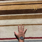 A close-up of a striped plush rug in beige, brown, and cream tones, with a person's hand placed on the surface to show scale. The brand name 'Atala Royal' is printed in gold near the top right.