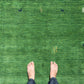 A top-down view of bare feet standing on a green textured rug with small embroidered motifs, including a corn cob and birds.