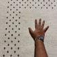 A close-up of a textured off-white carpet with a repeating dark brown tufted pattern, shown from above. A person's hand and forearm resting on the carpet provide a scale reference.