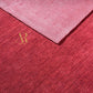 A close-up of a folded textured fabric or rug in two shades of pink-red: a lighter dusty rose on top and a deeper raspberry red below, with a small gold 'R' logo near the fold.