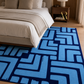 A rectangular area rug with a bold geometric maze-like pattern in bright blue and dark navy, styled on a light wood floor in a modern bedroom setting next to a bed.