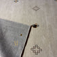 A close-up of a woven area rug corner showing the underside texture and a small tassel, placed on top of the same rug surface. The rug has a light beige top with subtle geometric motifs in brown, gray, and muted gold.