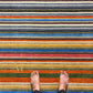 A top-down view of a person's bare feet standing on a thick, multicolored striped rug with horizontal bands of blue, red, orange, yellow, green, and gray.