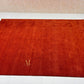 A rectangular red-orange rug with a subtle woven texture and a small gold monogram logo in the lower-left corner, displayed on a white floor against a beige textured wall.