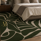 A green and beige abstract leaf-patterned area rug styled in a modern bedroom setting next to a bed with neutral bedding.