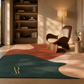 A large abstract area rug in a modern living room setting, featuring large organic shapes in dark teal, rust red, cream, and beige. The rug is styled in a warm, sunlit room with a wooden lounge chair, a floor lamp, and a bookshelf in the background.