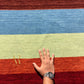 A rectangular area rug with vertical stripes in multiple colors including brown, blue, red, orange, pink, yellow, green, and light blue, featuring a gold embroidered monogram logo in the bottom left corner.