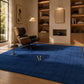 A rectangular royal blue textured rug with a small gold embroidered monogram logo in the upper right quadrant, shown flat against a plain white background.