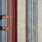 A top-down view of a striped wool rug with vertical bands of cream, light blue, burgundy red, and thin dark stripes. A person's bare feet and lower legs in black pants are visible on the left side of the rug, and a gold 'AR' logo watermark is visible on the right side.