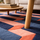 Colorful geometric-patterned rug with a wooden leg of a chair on it, in a living room setting.