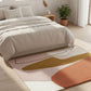 Neatly made bed with light bedding on a colorful abstract rug in a bright bedroom.