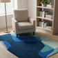 Modern living room with a beige armchair, geometric blue and green rug, and a bookshelf.